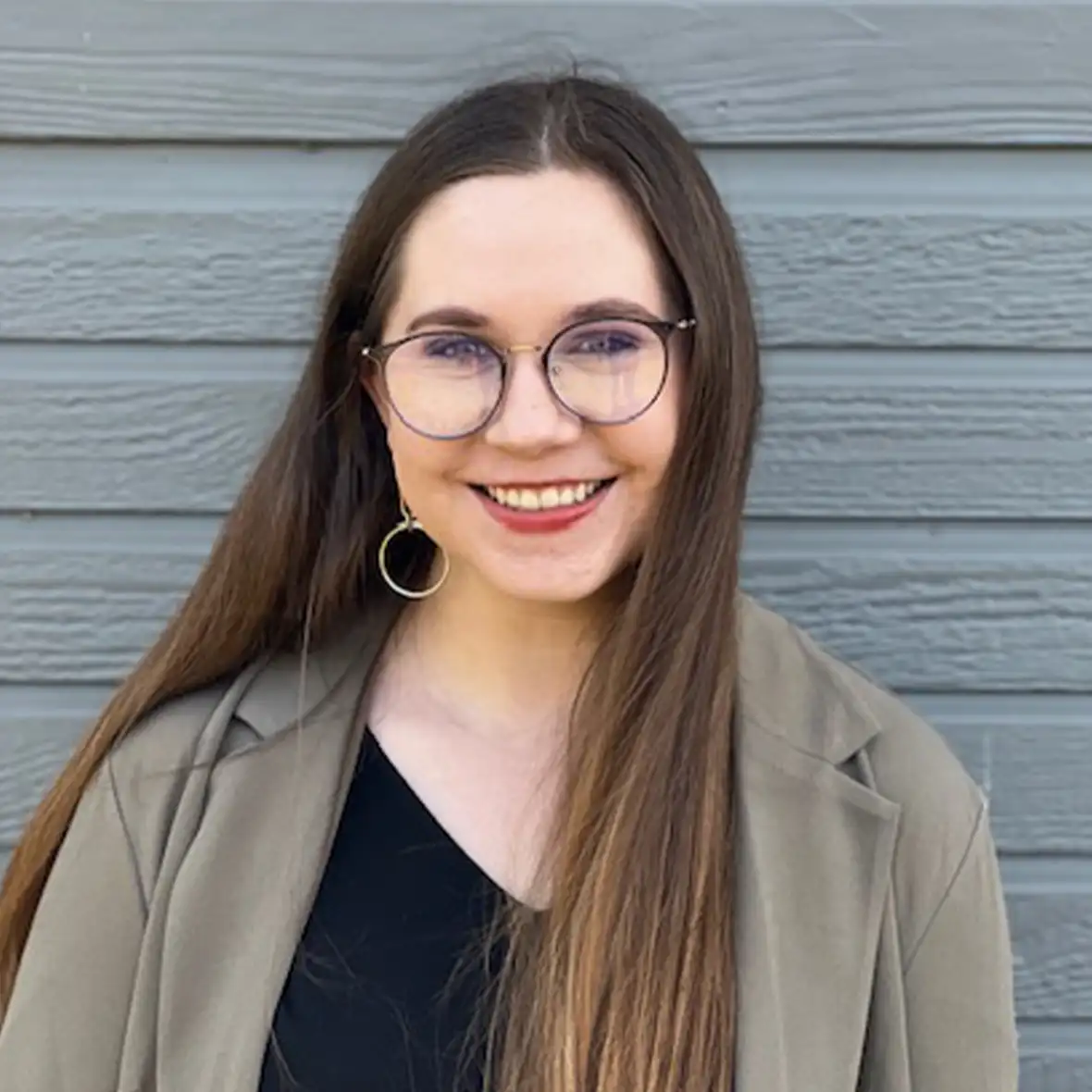 A person with glasses smiles in front of a gray wall, wearing a black top and beige jacket.