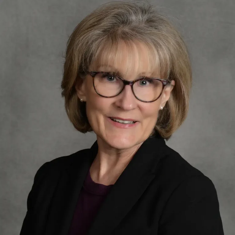 Woman with glasses and short hair wearing a black blazer, smiling at the camera.