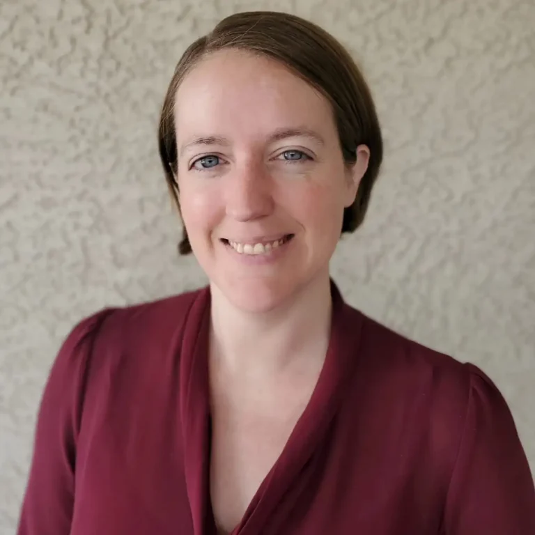 A person with short brown hair smiles against a textured wall background.