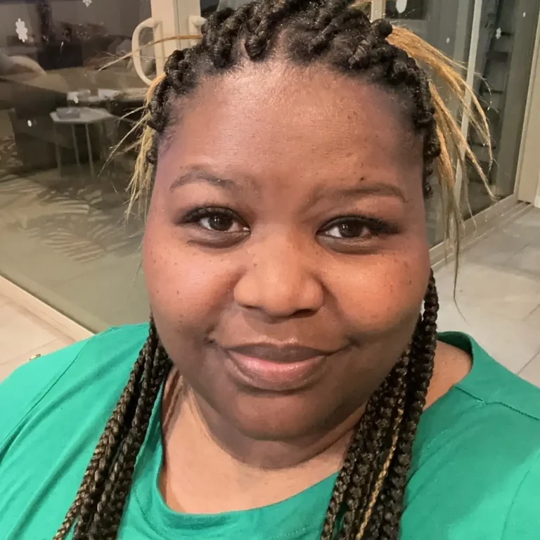 Person with braided hair wears a green top and smiles at the camera indoors.