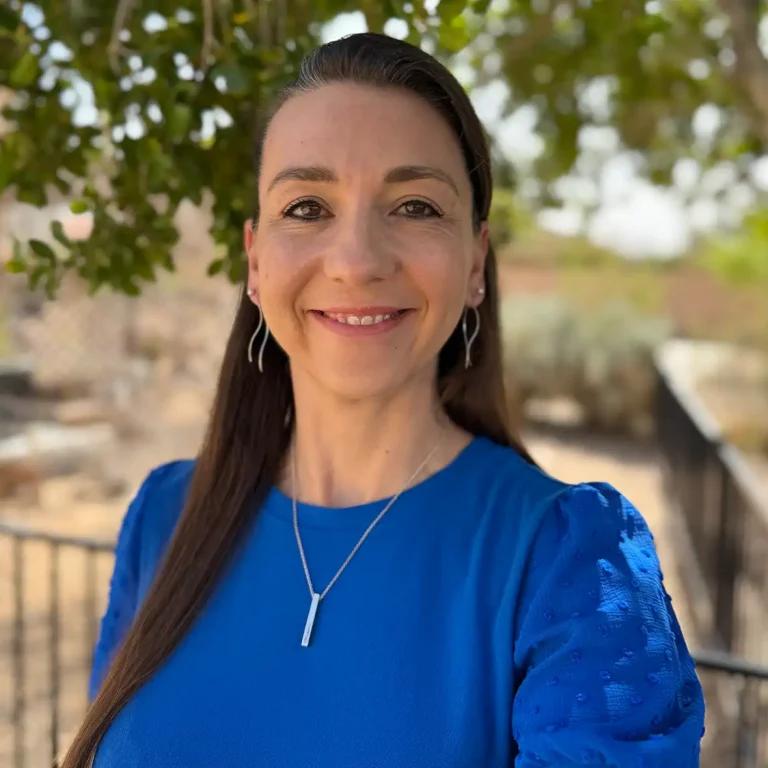Woman in a blue dress smiles, standing outdoors with trees in the background.